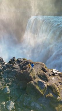 Ziyaretçiler Dettifoss şelalesinin kenarında, güçlü akıntıdan yükselen şiddetli şelaleye ve sise hayret ederek duruyorlar. Çarpıcı manzara İzlanda 'nın saf güzelliğini sergiliyor..