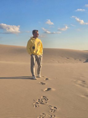 Genç bir adam Danimarka 'nın sakin kum tepelerinde geziniyor, yumuşak kumda ayak izleri bırakıyor. Parlak sarı kapüşonlu, kumulların sıcak tonlarıyla çok güzel zıtlaşıyor..