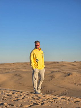 Bir gezgin, geniş kumlu tepelerin ortasında parlak sarı bir kapüşonlu giysiyle Danimarka 'nın çarpıcı manzarasının tadını çıkarıyor. Açık mavi gökyüzü, sahnenin sakin ve maceraperest atmosferini arttırır..