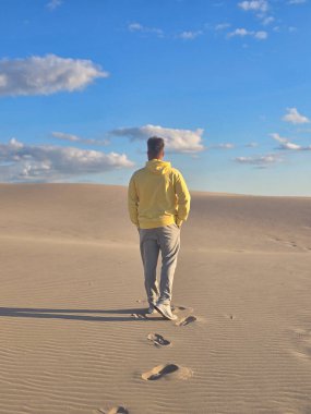Danimarka 'da yumuşak kum tepelerinde tek başına yürüyen bir gezgin, soluk kesici bir gökyüzü ile çevrili, tüylü bulutlarla dolu. Sakin atmosfer bir macera ve yansıma hissi uyandırıyor..