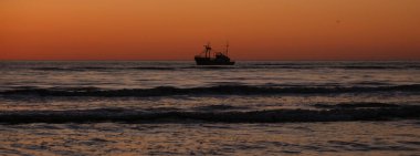 Bir balıkçı teknesinin titreyen dalgalar ve muhteşem bir günbatımı gökyüzü ile çevrili sakin denizde gezindiği sakin bir an. Danimarka 'da sahil güzelliği arayanlar için mükemmel..