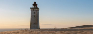 Günbatımının altın ışığında dimdik duran Rubjerg Knude Deniz feneri, Danimarka 'nın güzel, kayan kum tepeleriyle çevriliyken Kuzey Denizi üzerinde çarpıcı manzaralar sunuyor..