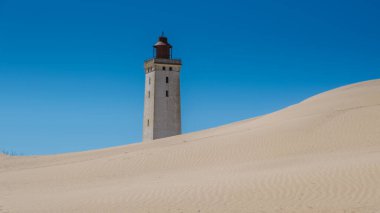 Zarif bir deniz feneri Rubjerg Knude Danimarka 'daki altın kum tepelerinden zarif bir şekilde yükseliyor, parlak mavi bir gökyüzü ile çevrili. Bu sakin kıyı manzarası huzurlu bir ortamda doğa güzelliğini sergiliyor..