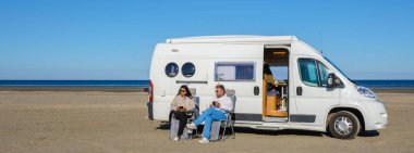 İki insan güzel bir Danimarka plajında huzurlu bir anın tadını çıkarıyor, rahat karavanlarının dışında içkilerini yudumluyorlar. Açık gökyüzü ve kumlu sahil rahatlama ve macera için mükemmel bir atmosfer yaratır..