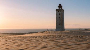 Altın güneş ışığı, Danimarka kıyıları yakınlarındaki kumullarda dimdik duran beyaz deniz fenerini yıkıyor. Huzurlu atmosfer doğanın güzelliğini ve Rubjerg Knude Danimarka 'nın sahil cazibesini yansıtıyor.