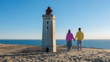 Danimarka 'da tarihi bir deniz fenerinin yanında kumlu kıyı şeridi boyunca el ele gezintiler. Güneş sakin denizin üzerinde ışıldıyor, Rubjerg Knude, Danimarka