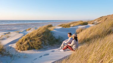 Güneş, Blokhus Strand, Danimarka 'daki sakin plajın üzerinde batarken, bir çift, yemyeşil çimlerle çevrili sıcak kum tepelerinde oturup, huzurlu anın tadını çıkarıyorlar..