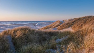 Danimarka kıyı şeridinde sakin bir gün batımı yumuşak kum tepeleri ve nazik dalgalar sergiliyor. Yakınlarda bir minibüs var, Vejers Caddesi.