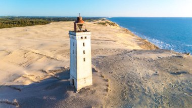 Danimarka 'da kumlu bir plaja kurulmuş tarihi bir deniz fenerinin çarpıcı manzarası. Güneş ufukta batıyor, kum tepeleri ve okyanus boyunca sıcak ışık saçıyor, Rubjerg Knude deniz feneri.