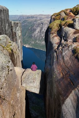 Tek bir figür, yüksek bir uçurumun tepesine tünemiş büyük bir kaya oluşumunun üzerinde oturur. Kjeragbolten Lysefjord, Norveç
