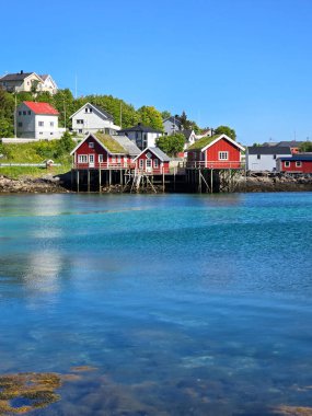 Tuhaf kırmızı kulübeler, açık mavi gökyüzünün altındaki pitoresk bir Norveç köyünün kıyı şeridini gösteriyor. Yansıtıcı sular doğanın güzelliğini besler, Lofoten Norveç