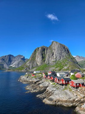 Açık mavi gökyüzünün altında dramatik dağlarla çevrili ikonik kırmızı kulübelerin bulunduğu Norveç fiyordunun nefes kesici görüntüsü. Lofoten Norveç