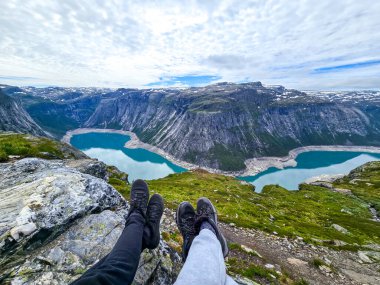 Engebeli bir uçurumdan nefes kesici manzaranın tadını çıkarırken, iki çift bacak kenardan sarkıyor. Aşağıda, turkuaz göller dramatik dağ zirvelerinin altında parıldıyor. Norveç 'te Trolltunga yürüyüşü