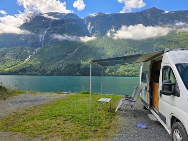 Rahat bir karavan, sakin bir gölün yanına park edilmiş. Arka planda nefes kesici dağlar var. Rahatlatıcı sandalyeler ziyaretçileri Norveç doğasının dingin güzelliğine davet ediyor..