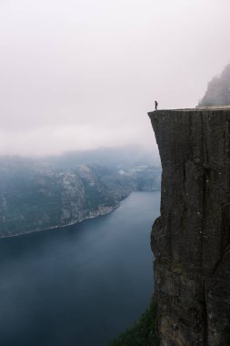 Yalnız bir figür yükselen bir uçurumun kenarında duruyor, Preikestolen Norveç 'in dramatik fiyortlarına bakıyor. Yoğun sis dağları sarıyor, macera ve korku hissini arttırıyor..