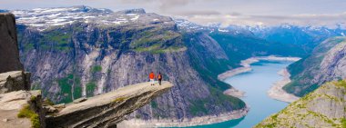 İki yürüyüşçü, Trolltunga 'ya, Norveç' in görkemli fiyortlarına ve yemyeşil vadilerine bakan dramatik bir uçurum kenarında duruyor. Gökyüzü parçalı bulutlu, bu büyüleyici manzara için mükemmel bir zemin oluşturuyor..