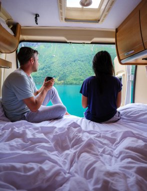 İki insan rahat bir karavanda rahatlar, kahveyi yudumlarken nefes kesici Norveç manzarasına hayran kalırlar. Sabahın erken saatlerinde yemyeşil dağlar ve sakin sular, huzur ve macera hissi uyandırır..