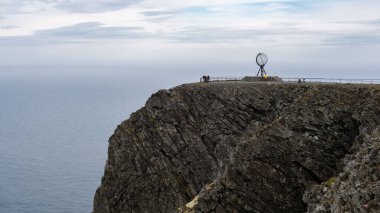 Ziyaretçiler Nordkapps 'taki dramatik kayalıklarda toplanarak engin okyanusa ve bulutlu bir gökyüzünün altındaki engebeli araziye bakıyorlar. Bu ikonik konum nefes kesici manzaralar ve macera hissi sunuyor..
