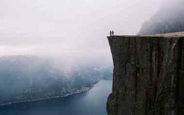 Preikestolen Norveç 'teki yüksek bir uçurumun tepesinde duran iki maceraperest, aşağıdaki muhteşem fiyorda bakıyor, ruhani sisle çevrili. Doğanın ve keşiflerin nefes kesici bir anı..