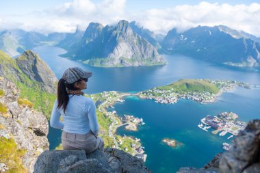 Bir kadın, Reine köyünün ve Lofoten Norveç 'in etrafındaki fiyortların çarpıcı panoramik manzarasının keyfini çıkarıyor. Görkemli dağlar kristal berraklığında suların üzerinde yükselir, dingin ve görkemli bir manzara yaratır..