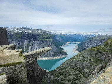 Norveç 'te muhteşem bir fiyordun üzerinde yükselen görkemli kayalıklar zümrüt sularının panoramik manzarasını sunuyor. Kenarda yalnız bir yürüyüşçü duruyor, Trolltunga Norveç yürüyüşü