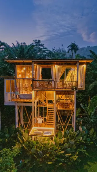 Yeşilliğe gömülü Bali 'deki bu göz kamaştırıcı ağaç ev alacakaranlıkta huzurlu bir kaçış sunuyor. Misafirler güvertede dinleniyor, yağmur ormanlarının güzelliğiyle çevrili, yumuşak ışıklarla aydınlatılmış..