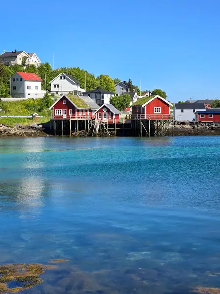 Tuhaf kırmızı kulübeler, açık mavi gökyüzünün altındaki pitoresk bir Norveç köyünün kıyı şeridini gösteriyor. Yansıtıcı sular doğanın güzelliğini besler, Lofoten Norveç
