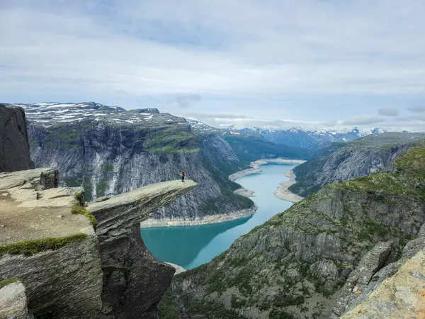 Norveç 'te muhteşem bir fiyordun üzerinde yükselen görkemli kayalıklar zümrüt sularının panoramik manzarasını sunuyor. Kenarda yalnız bir yürüyüşçü duruyor, Trolltunga Norveç yürüyüşü