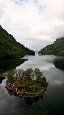 Bereketli yeşillik ve sakin yansıtıcı sularla çevrili büyüleyici kırmızı bir kulübenin bulunduğu sakin bir Norveç adasının büyüleyici manzarası. Lovrafjorden, Norveç