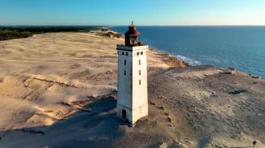 Danimarka 'nın Jutland kentindeki Rubjerg Knude Fyr Deniz Feneri' nin nefes kesici güzelliğini keşfedin. Altın kumlar, hafif dalgalar ve canlı bir günbatımı gökyüzüne karşı görkemli bir deniz feneri., 