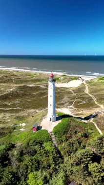 Lyngvig Deniz Feneri Hvide Sande 'nin Jutland, Danimarka' daki nefes kesici güzelliğini keşfedin. Açık mavi gökyüzü altında, kıyı manzaraları ve kumlu sahillerin ortasındaki ikonik deniz fenerini gösterin..