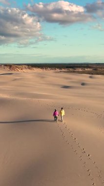 Danimarka, Jutland 'in altın kum tepelerinde el ele yürüyen iki insan güneş batarken yumuşak yüzeyde ayak izleri yaratıyor Rabjerg Mile kum tepeleri.