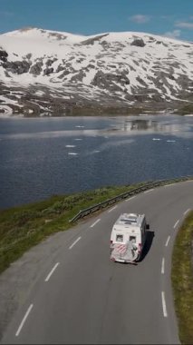 Norveç 'te karla kaplı görkemli dağlarla çevrili sakin bir göl kenarındaki dolambaçlı bir yolda bir karavan seyahat ediyor. Açık mavi gökyüzü hayranlık uyandıran manzarayı tamamlıyor..