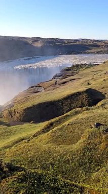 Görkemli İzlanda arazileri buzul nehirlerine bakan yemyeşil tepeleri sergiliyor. Mekan, açık mavi gökyüzü altında doğanın saf güzelliğini yansıtıyor, Dettifoss şelalesi İzlanda.