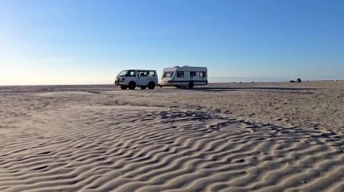 Romo Lakolk plajında huzurlu bir akşam yumuşak, kumlu sahilde park etmiş bir karavan sergileniyor. Sakin atmosfer ve geniş gökyüzü plaj aşıkları ve gezginler için pitoresk bir geri çekilme yaratıyor..