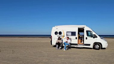 İki kişi, karavanlarının önünde oturan Romo Lakolk Plajı 'nda huzurlu bir günün tadını çıkarıyor. Uçsuz bucaksız kumlu sahil ve sakin okyanus dinlenmek ve keşfetmek için mükemmel sakin bir atmosfer yaratır..