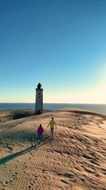 Rubjerg Knude Fyr Deniz Feneri, Jutland 'in muhteşem kumlu sahilinde, canlı akşam gökyüzünün altında el ele bir yürüyüş. Uzak deniz feneri huzurlu manzaraya cazibe katar..