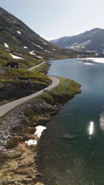 Norveç 'te yemyeşil ve görkemli karlı zirvelerle çevrili sakin bir gölün kenarını saran dolambaçlı bir yolun nefes kesici manzarası. Doğa ve seyahat meraklıları için mükemmel..