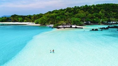 İki insan sığ turkuaz sularda yürürken yumuşak dalgalar yavaşça kıyıya çarpar. Koh Mak Tayland yakınlarındaki Koh Kham Adası.