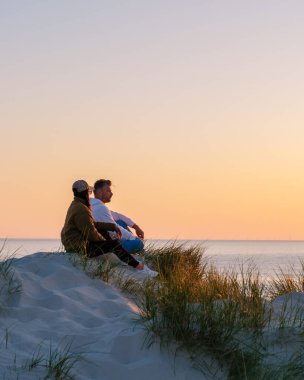İki arkadaş kumlu bir kumulda Danimarka sahilinin sakin sularında gün batımını seyrederek rahatlıyor. Gökyüzünün sıcak renkleri resim gibi bir zemin oluşturur, Vejers Strand Sahili Danimarka