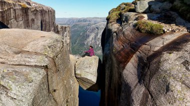 Tek başına bir yürüyüşçü, Norveç 'in Kjeragbolten Lysefjord kayalıklarına bakan devasa bir kayanın üzerinde oturur. Açık mavi gökyüzü ve dramatik manzara mükemmel bir yürüyüş deneyimi yaratır..