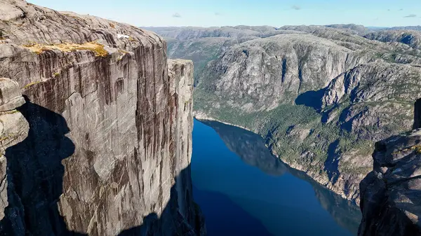 Görkemli uçurumlar Norveç 'teki sakin bir fiyordun üzerinde belirgin bir şekilde yükselirler, açık mavi gökyüzünü yansıtırlar. Bu büyüleyici manzara macera arayıcılarını davet ediyor, Kjeragbolten Lysefjord, Norveç