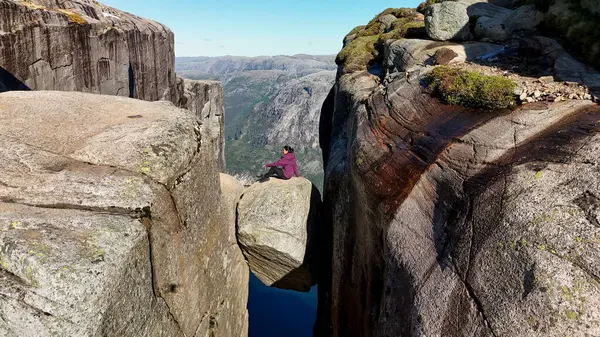 Tek başına bir yürüyüşçü, Norveç 'in Kjeragbolten Lysefjord kayalıklarına bakan devasa bir kayanın üzerinde oturur. Açık mavi gökyüzü ve dramatik manzara mükemmel bir yürüyüş deneyimi yaratır..