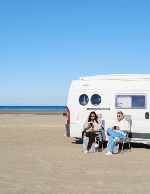 İki gezgin, Lakolk Strand, Romo Danimarka 'da kumlu bir plajda karavanın önünde otururken atıştırmanın tadını çıkarıyorlar. Açık mavi gökyüzü ve sakin deniz bir günlük rahatlama için mükemmel bir zemin oluşturur..
