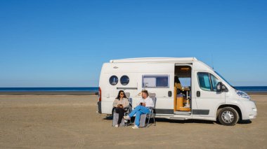 Arkadaşlar plajda huzurlu bir günün tadını çıkarırlar, karavanın dışında dinlenirler. Açık mavi gökyüzü ve sakin deniz rahatlık ve dostluk için mükemmel bir zemin sağlar. Lakolk Strand, Romo Danimarka