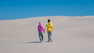 Renkli kapüşonlu giyinmiş genç çift kumlu kumulların arasında el ele geziyor parlak mavi gökyüzüne karşı. Huzurlu atmosfer, Danimarka 'nın Rabjerg Mile kumullarında keşif yapmak için mükemmel bir günü yansıtıyor.