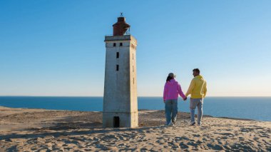 Kumlu bir sahil boyunca el ele yürüyüp açık mavi gökyüzünün altındaki Anholt Deniz Feneri manzarasının tadını çıkarıyorlar. Güneş ufukta batıyor, Rubjerg Knude, Danimarka