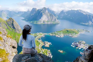 Büyüleyici manzara, Lofoten Norveç 'teki büyüleyici fiyortlara ve canlı köye hayran bir kadının bir kayanın üzerinde oturmasını içeriyor. Yeşillik, parçalı bulutlu gökyüzünün altındaki görkemli dağlarla çelişiyor..