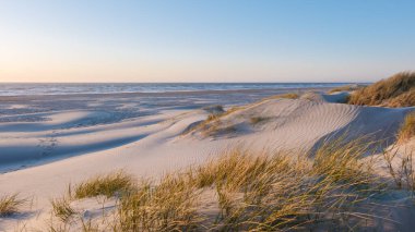 Yumuşak kum tepeciklerinin dingin okyanus dalgalarıyla canlı bir günbatımı gökyüzü altında buluştuğu Danimarka, Vejers Strand 'in dingin güzelliğini deneyimleyin.. 