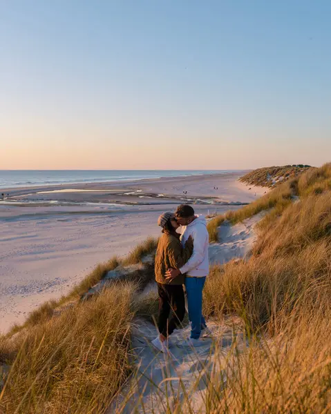 Danimarka 'da kumlu bir sahilde, canlı bir günbatımı gökyüzünün altında yemyeşil kumullarla çevrili bir çift öpüşüyor. Bu an deniz kıyısında sevgi ve sükuneti yakalar. Blokhus Strand Sahili